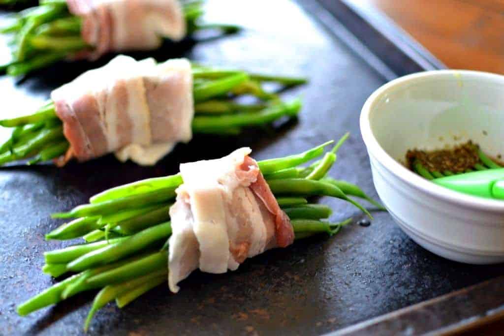 Green bean bundles wrapped in bacon on a baking sheet with a bowl of oil and seasonings on the side. 