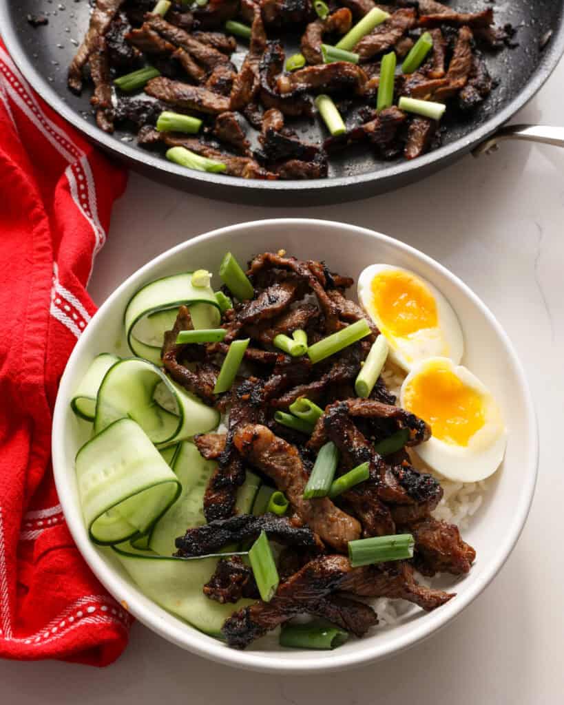 A single serving white bowl full of white rice, beef bulgogi, pickled cucumbers, and a soft boiled egg. 