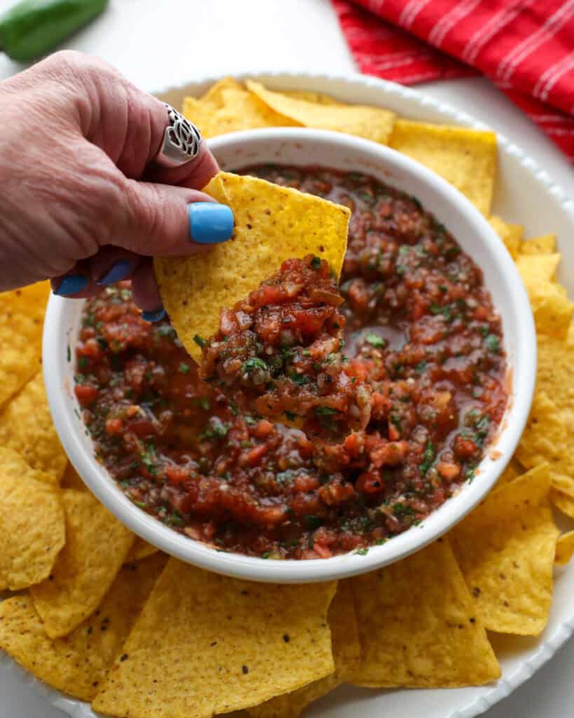 5 Minute Mouthwatering Fresh Salsa Recipe A chip full of tomato salsa over a bowl of salsa.