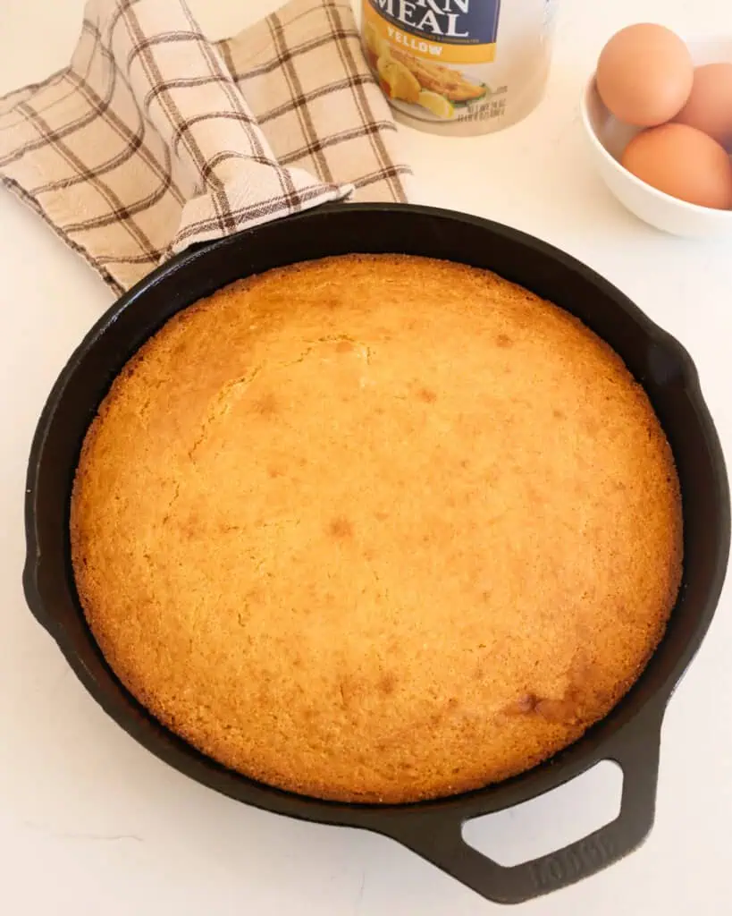 Fresh baked cornbread in a skillet with eggs and cornmeal in the background. 