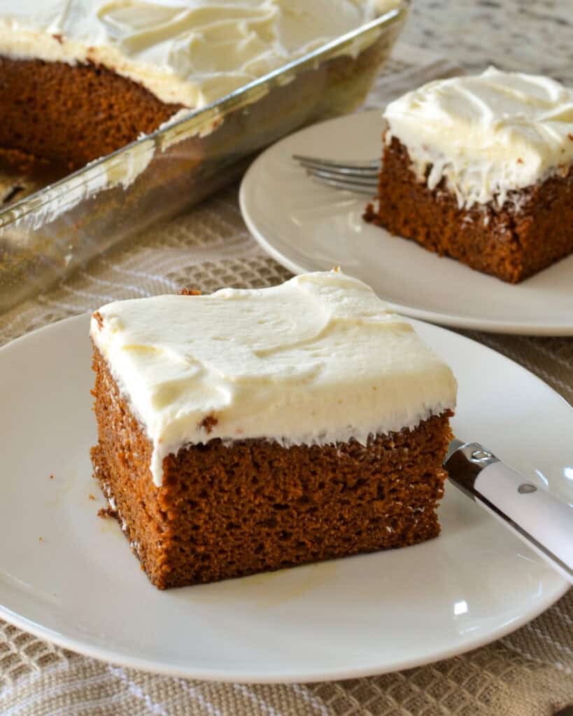 Gingerbread Cake A slice of gingerbread cake with cream frosting on a single serving plate with a fork with the gingerbread cakein the background.