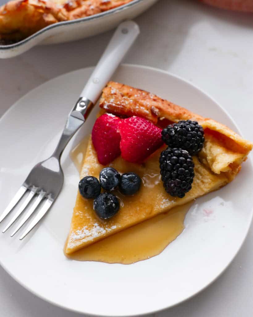 A piece of Dutch baby pancake with berries and warm maple syrup on a single serving plate with a fork. 