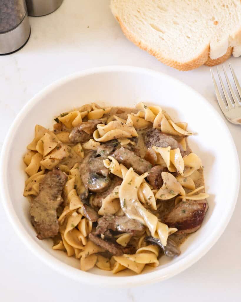 Slow Cooker Beef Stroganoff Beef stroganoff in a single serving bowl with bread and salt and pepper shaker in the background.