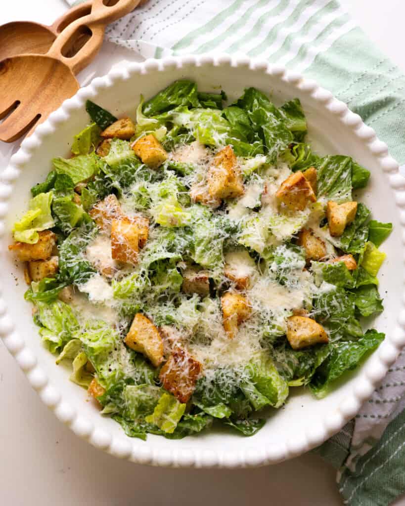 A large overhead view of a chicken Caesar salad in a white serving bowl with tongs in the background. 