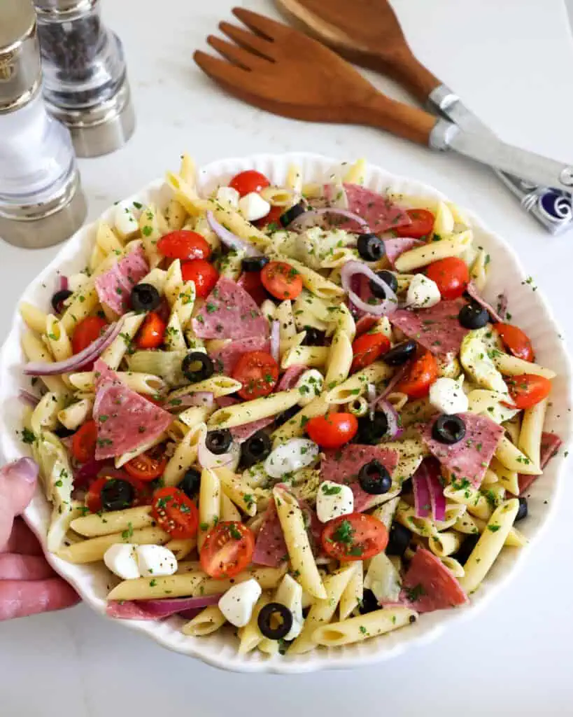 A large white serving bowl full of Italian pasta salad with serving utensils and salt and pepper shaker in the background. 