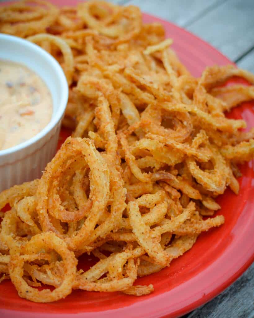 A close up view of onion straws on a red serving plate with dipping sauce. 