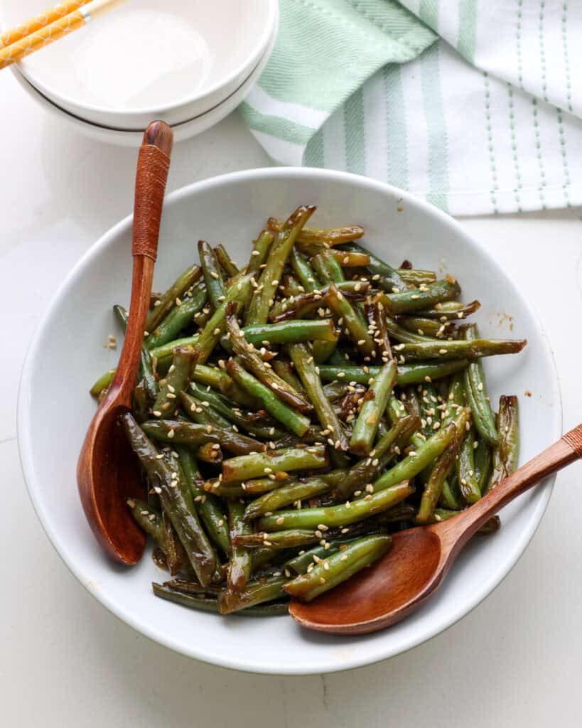 Stir Fried Green Beans A close up view of white serving bowl and two wooden spoons full of stir fried green beans with a green and white towel in the background.