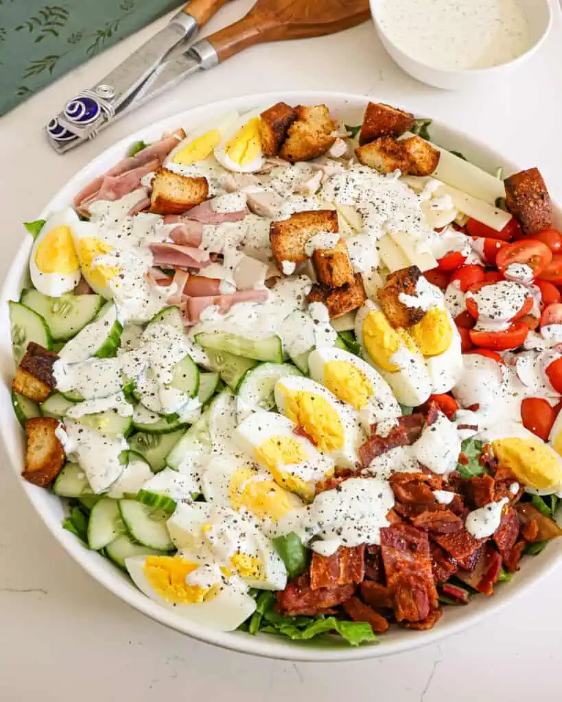 Chef Salad An overehead view of a chef salad with ranch dressing in a large bowl with a bowl of ranch dressing and serving utensils in the back ground.