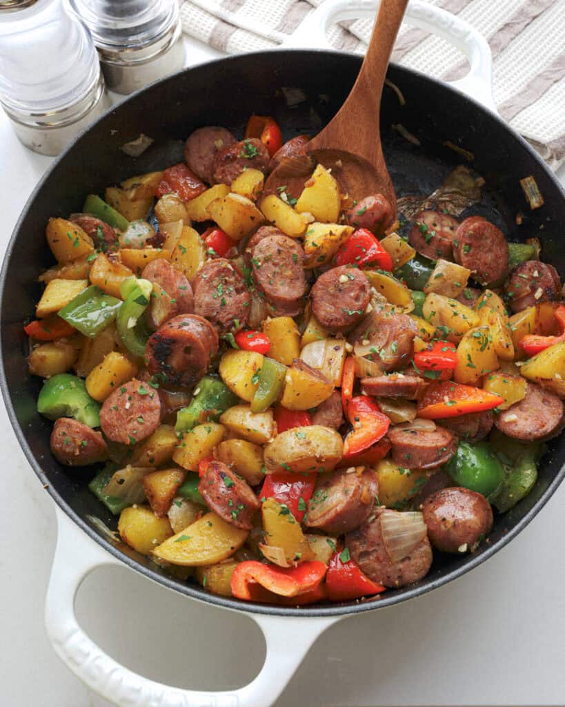 A large white cast iron skillet with potatoes, sausage, onions, and green and red bell pepper in it. 