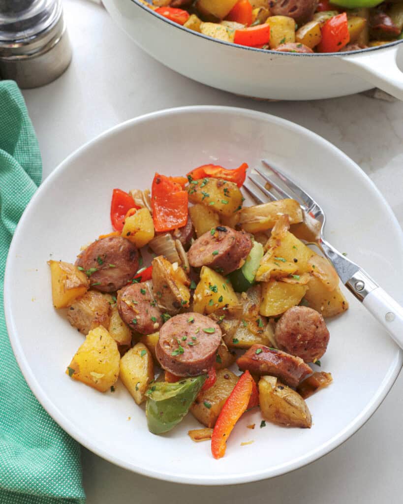 A single white serving bowl full of sausage and potatoes with rest of the skillet in the background. 