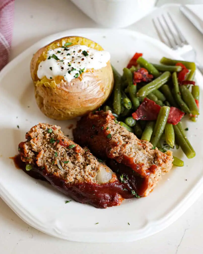 Moist Turkey Meatloaf A close up view of two slices of ground turkey meatloaf on a white serving plate with green beans and potatoes.
