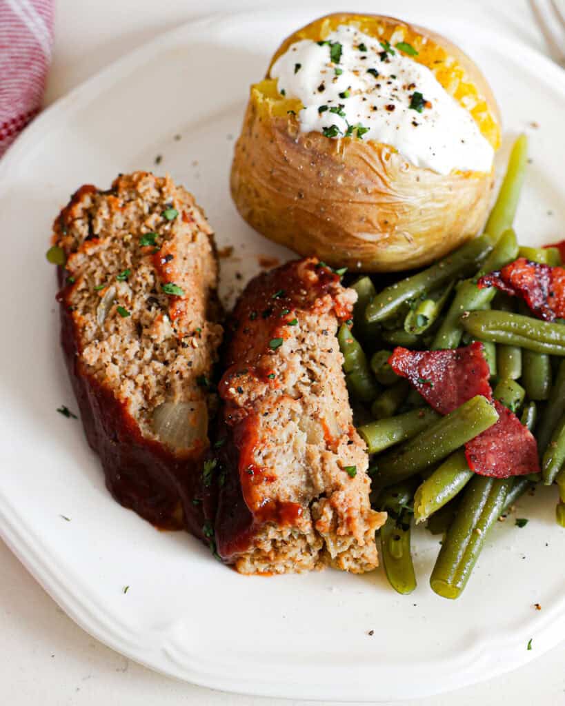 A single serving white plate with two slices of turkey meatloaf, green beans with bacon, and a baked potato with sour cream and chives. 