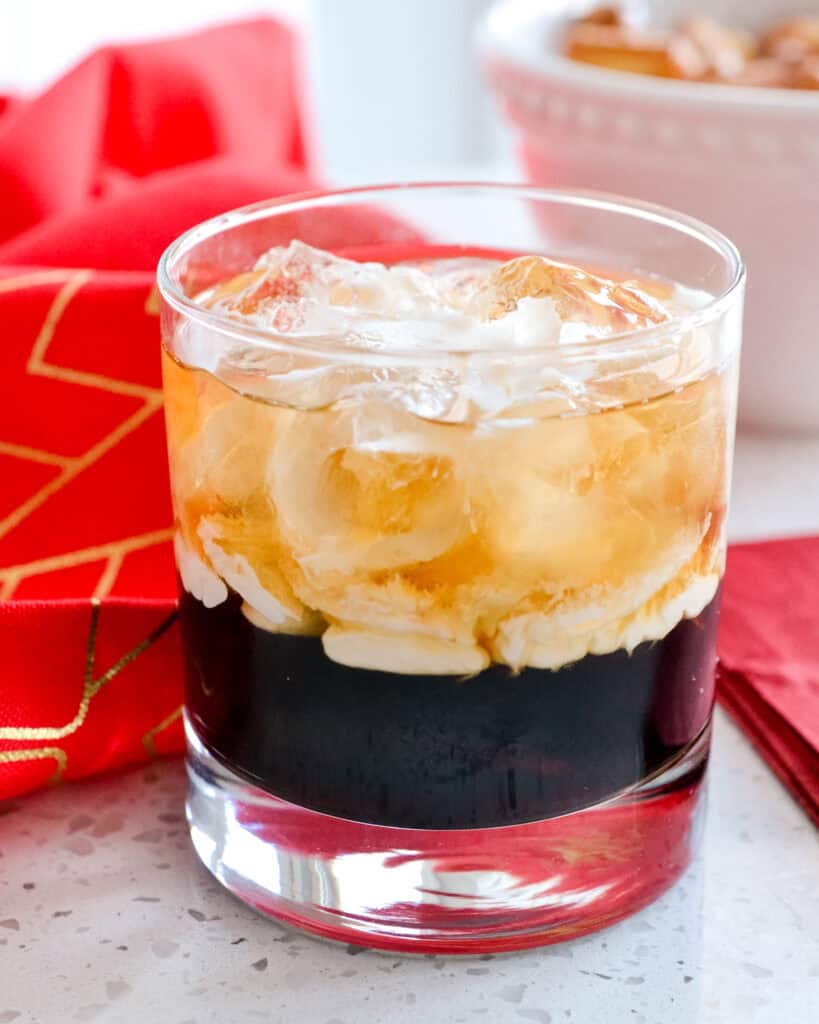 A white Russian cocktail in an old fashioned glass in front of a red towel and a bowl of pretzels. 