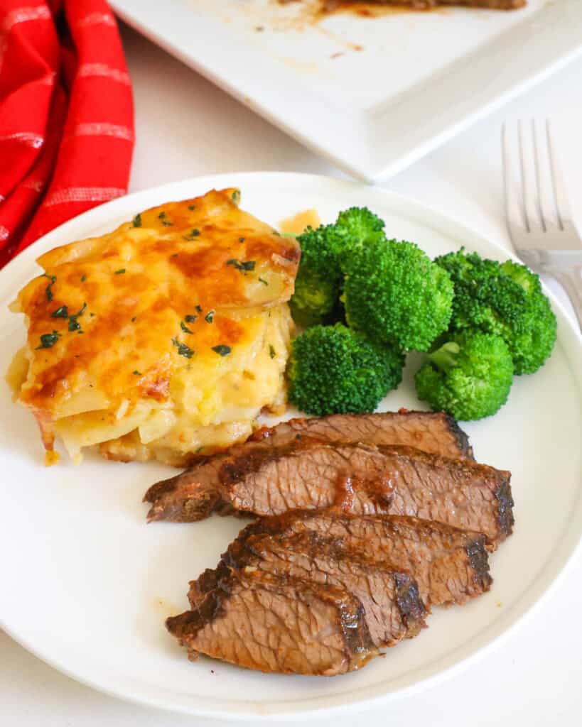 Sliced beef brisket on a serving plate with steamed broccoli and au gratin potatoes. 