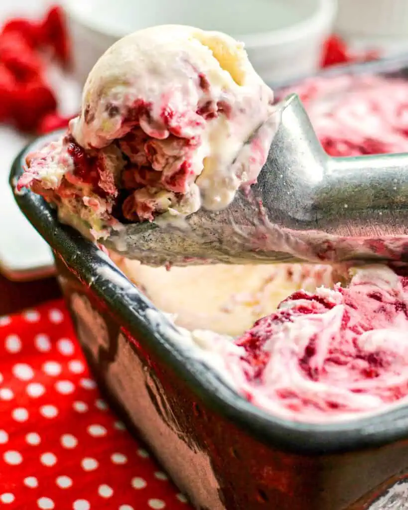 An ice cream sccop full of raspberry ice cream over a loaf pan full of raspberry ice cream. 