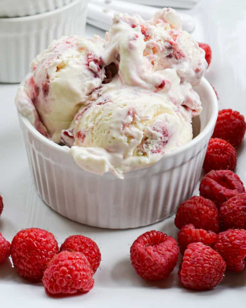 A white bowl full of raspberry ice cream on a plate with raspberries.