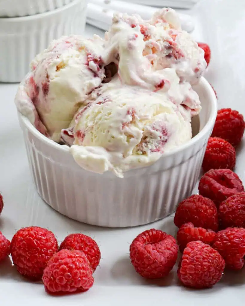 A white bowl full of raspberry ice cream on a plate with raspberries. 