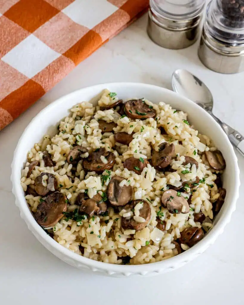 A serving bowl full of mushroom risotto with a spoon on the side and a kitchen towel in the background. 