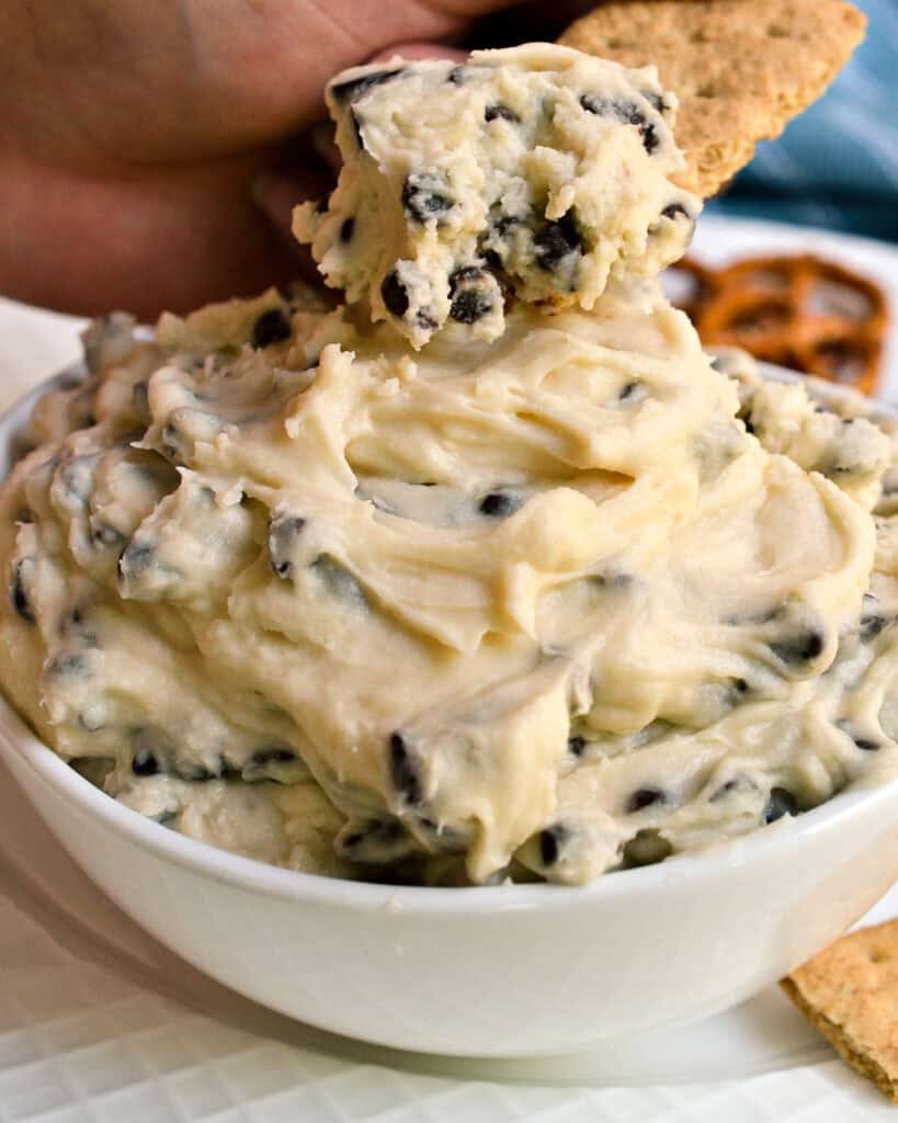 A white serving bowl of cookie dough dip with a graham cracker dipped in it.