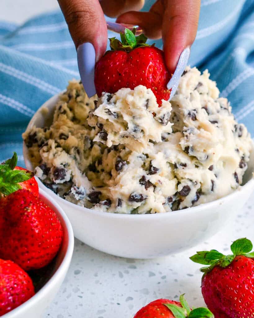 A bowl full of cookie dough dip with a strawberry dipped in it and an extra bowl of strawberries on the side.