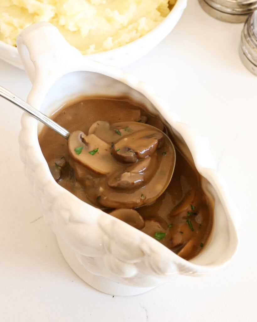 A gravy boat and ladle full of mushroom gravy with a salt and pepper shakter and a bowl of mashed potatoes in the background.