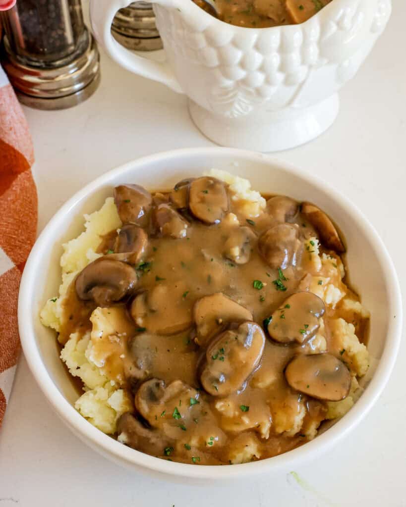 A bowl full of mashed potatoes smothered in muhsroom gravy with extra gravy in the gravy boat in the background with a salt and pepper shaker.