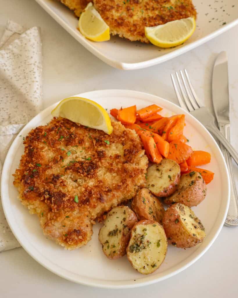 Pork schnitzel on a plate with roasted red potatoes and honey glazed carrots with a serving platter full of schnitzel in the background. 