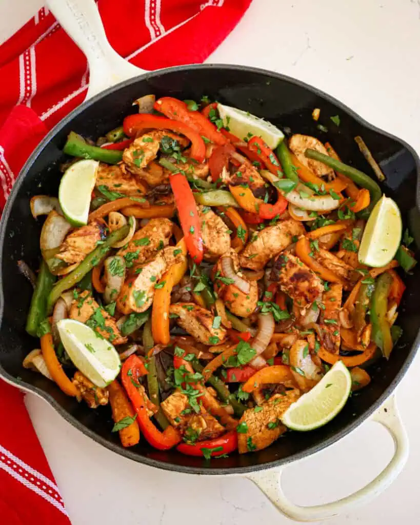 Overhead view of chicken fajitas in a large skillet with lime wedges and chopped cilantro. 