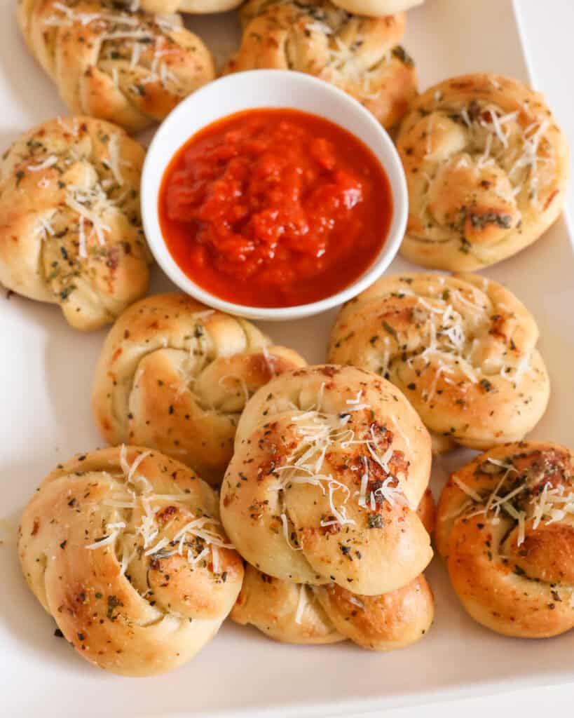 A white serving platter full of garlic knots and a bowl full of warm marinara.