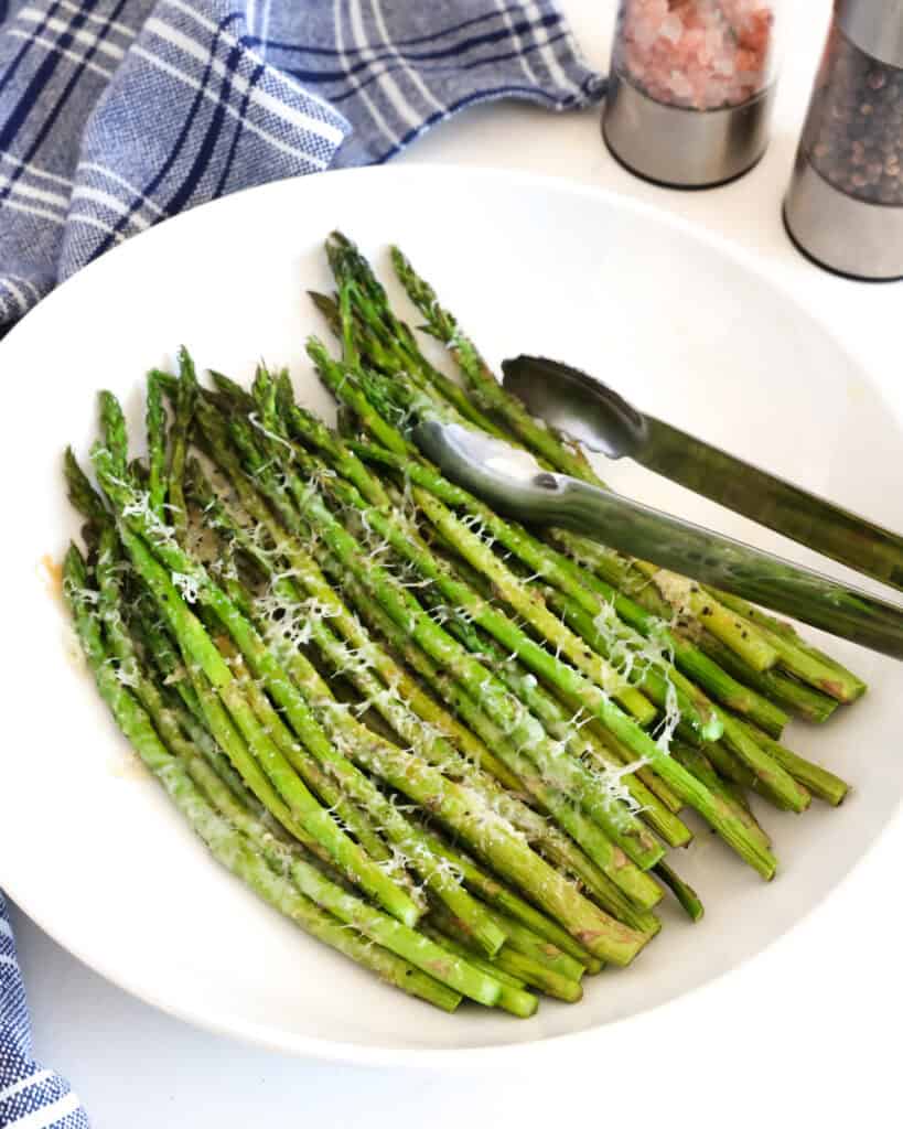 Air Fryer asparagus in a bowl with a salt and pepper shaker in the background and a blue towel in the background. 