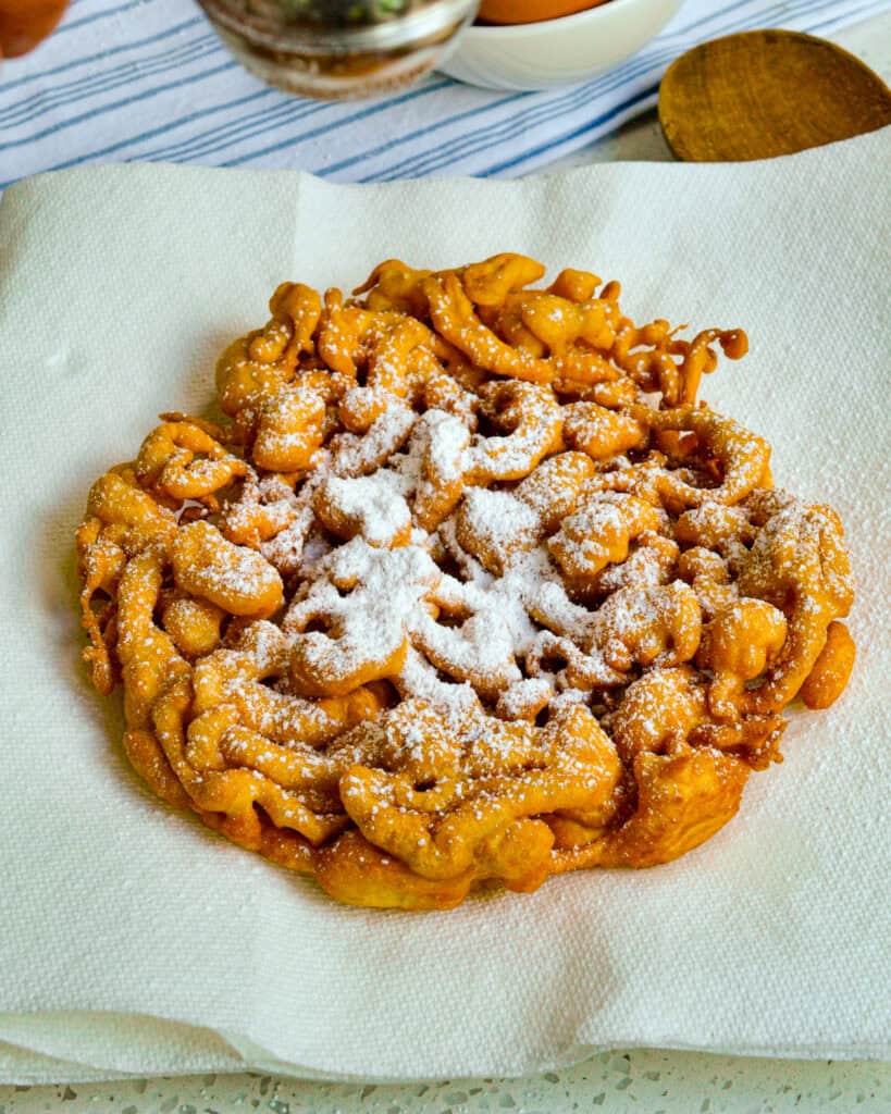 A funnel cake on a paper towel with powdered sugar sprinkled on it. 