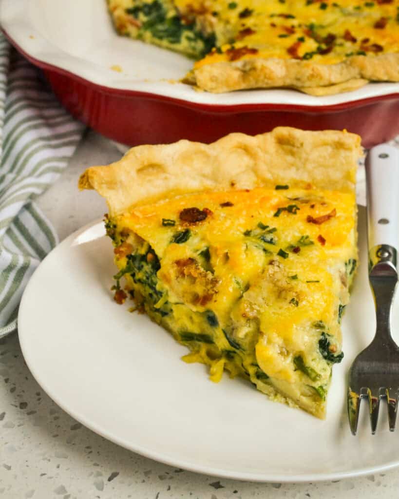 A close up view of a slice of spinach quiche on a plate with a fork and a green and white kitchen towel in the background. 