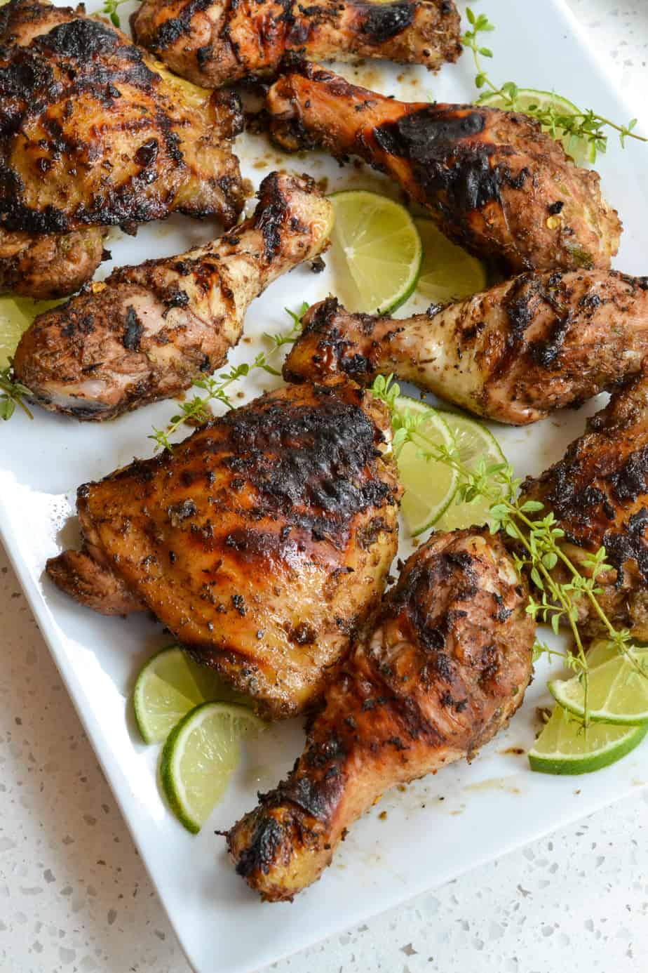 Grilled jerk chicken on a platter with lime slices and thyme. 