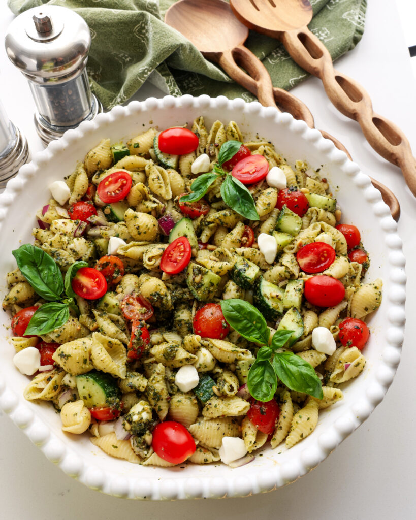 A big white bowl full of pesto pasta salad with tongs and a salt and pepper shaker in the background. 
