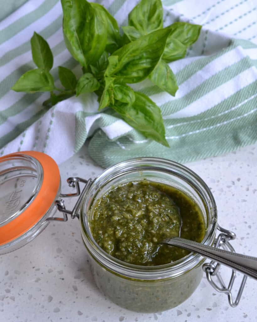 Pesto Sauce in a small jar with a spoon and fresh basil behind.
