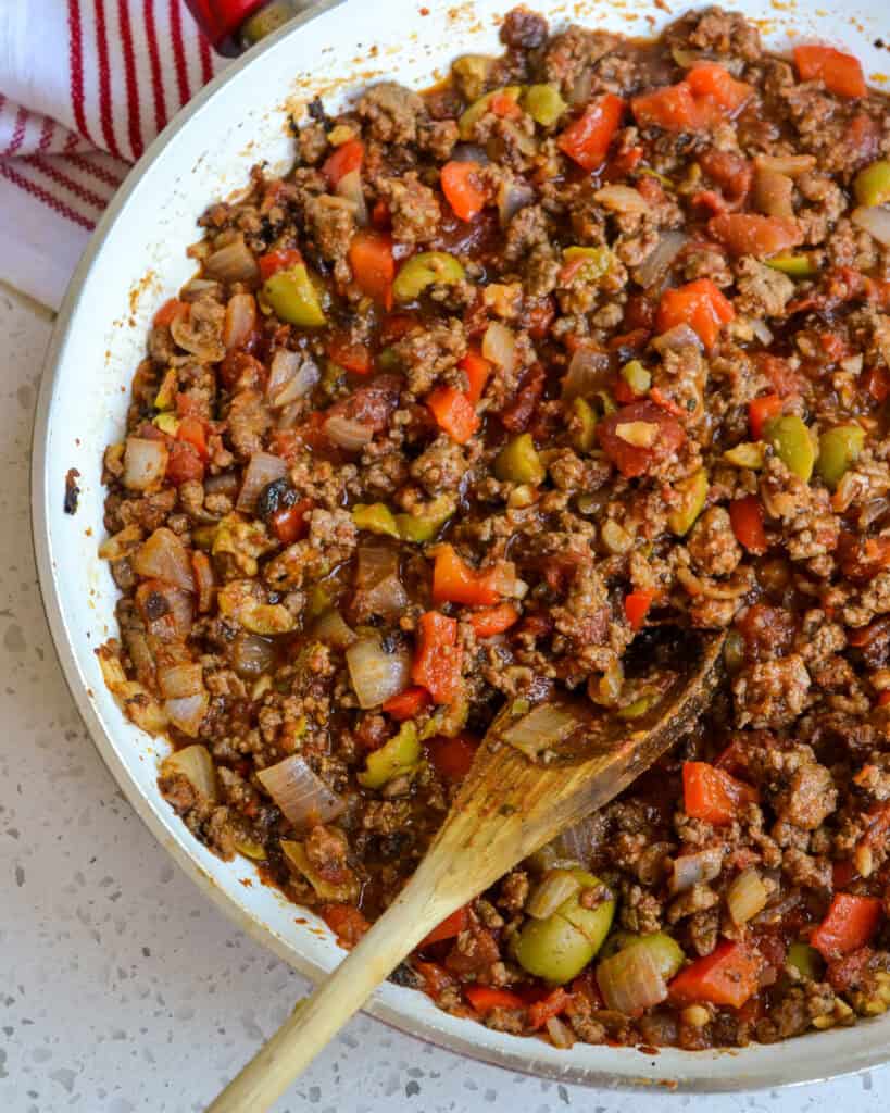 A skillet full of ground beef, tomatoes, tomato sauce, green olives, and chopped raisins.