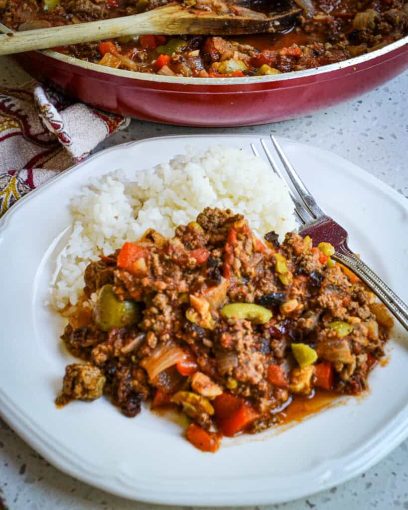 A plate full of Cuban Picadillo and white rice with a skillet full of picadillo in the background.