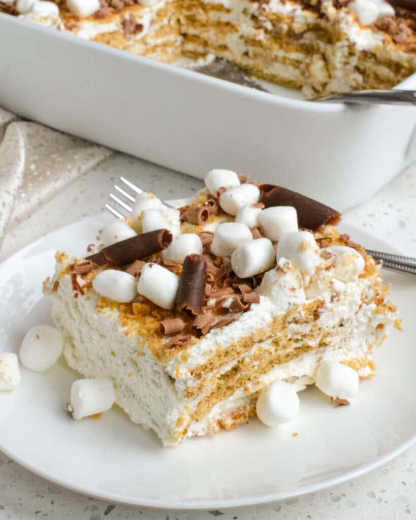 A slice of Icebox Cake topped with crushed graham crackers, mini marshmallows, and chocolate curls on a white plate.