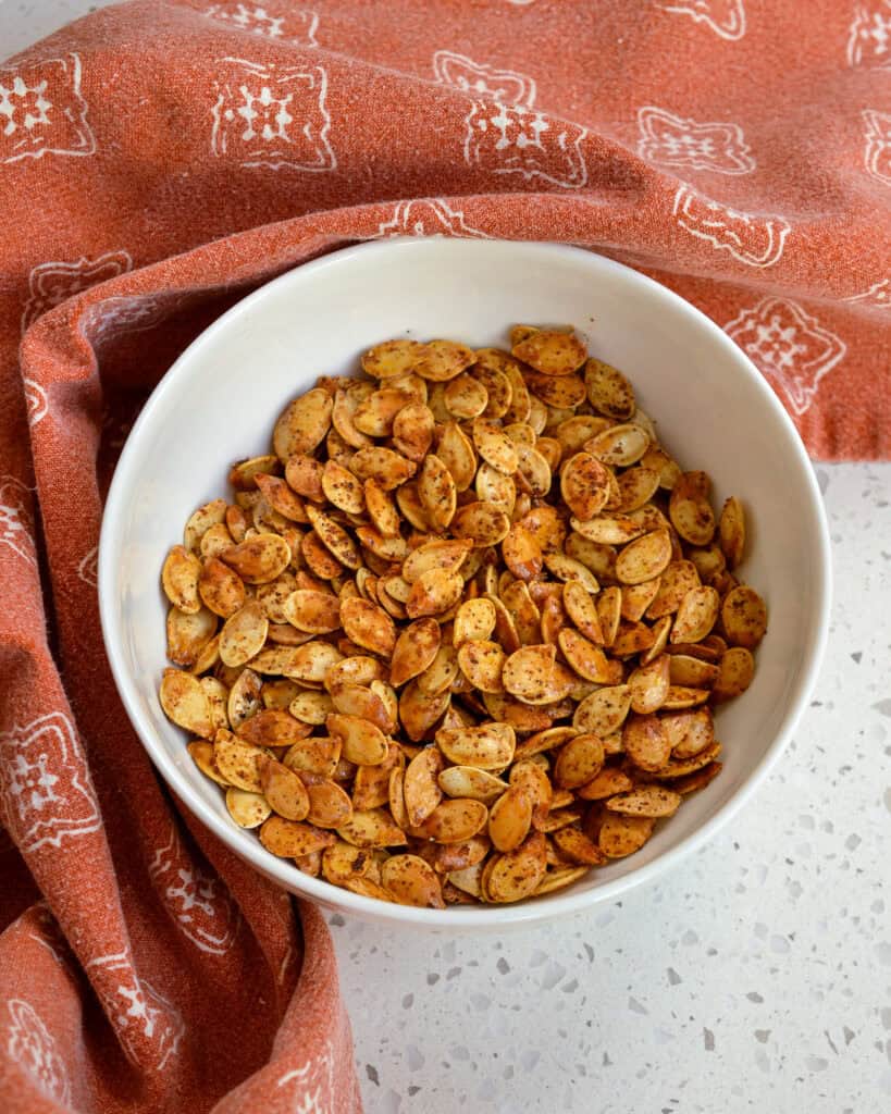 Roasted Pumpkin Seeds in a white bowl with an orange towel beside it.