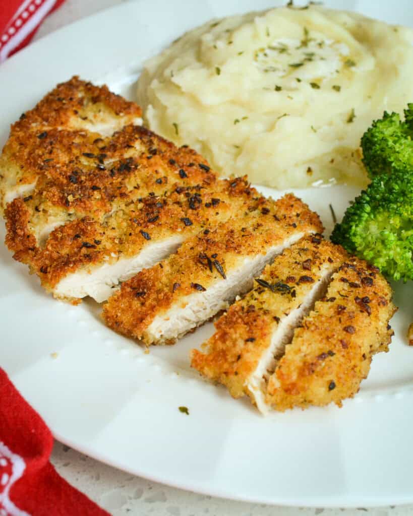 Parmesan Crusted Chicken cut into slices served on a white plate with mashed potatoes and broccoli.