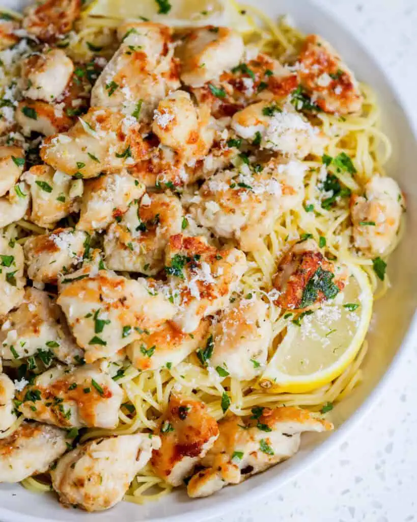 A close up view of chicken scampi in a serving bowl over thin spaghetti. It is garnished with Parmesan cheese, lemons, and chopped fresh parsley. 