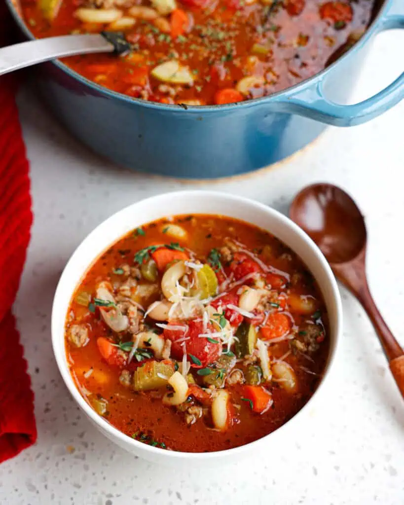 A single bowl full of Italian sausage soup with a wooden spoon and a Dutch oven full of the soup in the background.