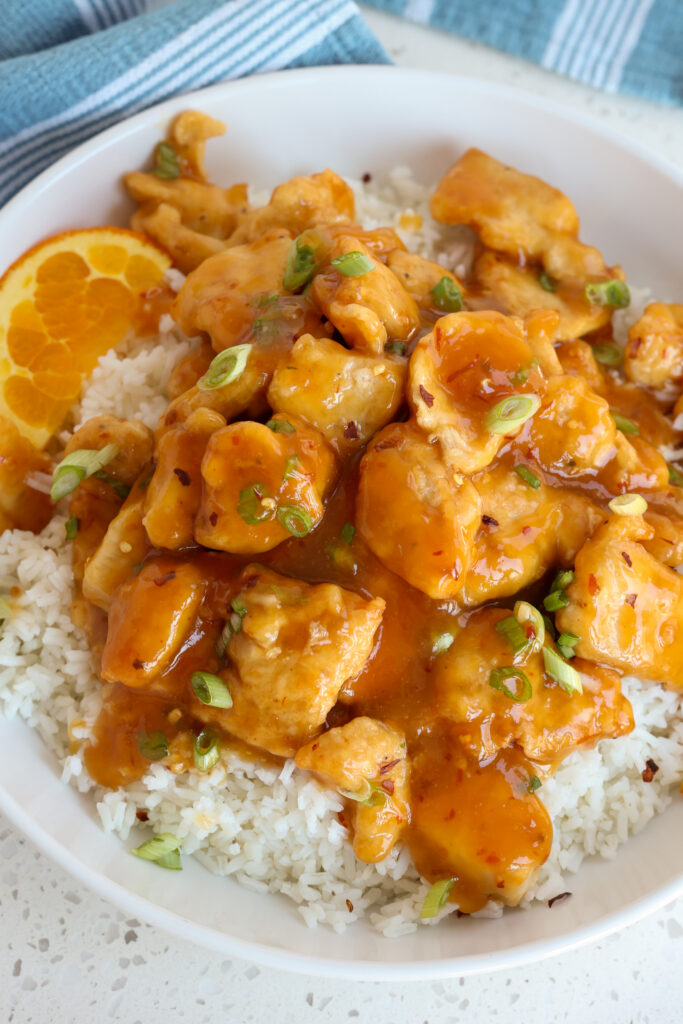 A bowl full of Orange Chicken with sauce, green onions, and crushed red pepper. 