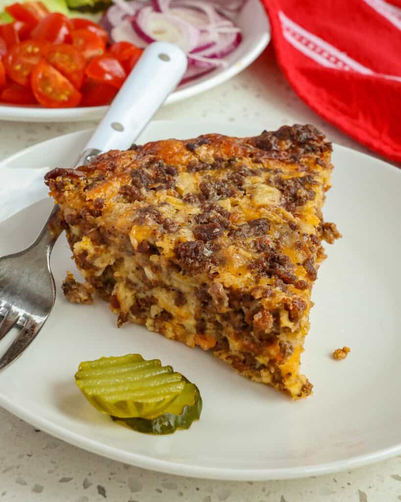 A slice of cheese cheeseburger pie on a single serving white plate with pickles and a fork. 