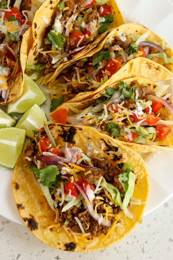 Four ground beef tacos on a plate with lettuce. tomato, red onions. and cheddar cheese