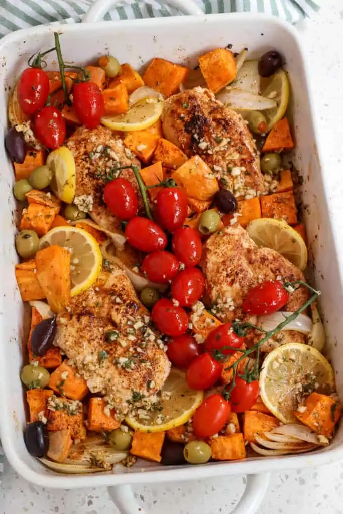 A tasty Greek Chicken Recipe with roasted onions, sweet potatoes, chicken breasts, cherry tomatoes, olives, and lemon all drizzled with a rosemary, lemon, and paprika marinade and topped with crumbled feta.