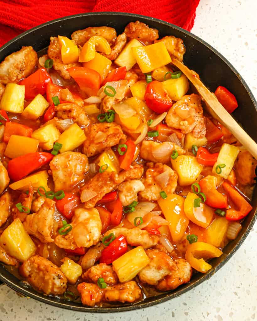 An overhead of pineapple chicken and a wooden spoon in a black skillet. 