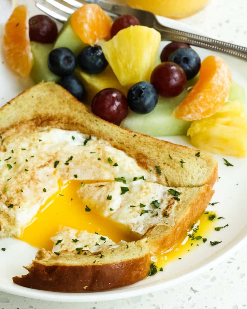Egg in a Hole cut open with fresh herbs on top and fresh mixed fruit on the side.