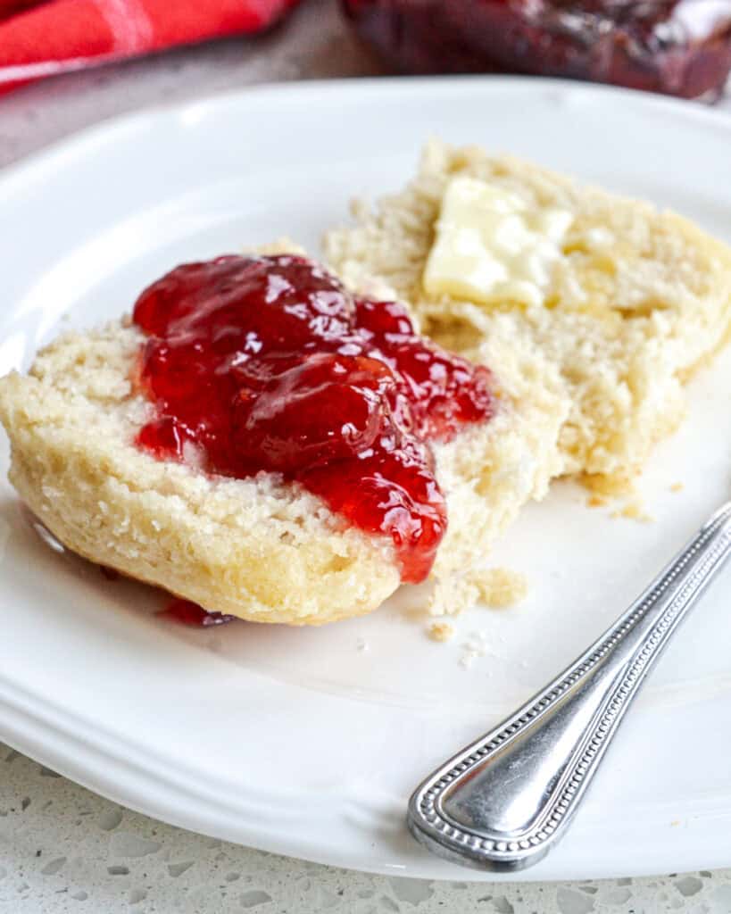 A split 7up biscuit with strawberry jam and butter on a white serving plate with a knife. 