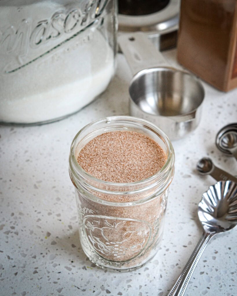 A small mason jar full of cinnamon sugar with a spoon, measuring cup, measuring spoons, granulated sugar, and ground cinnamon in the background. 